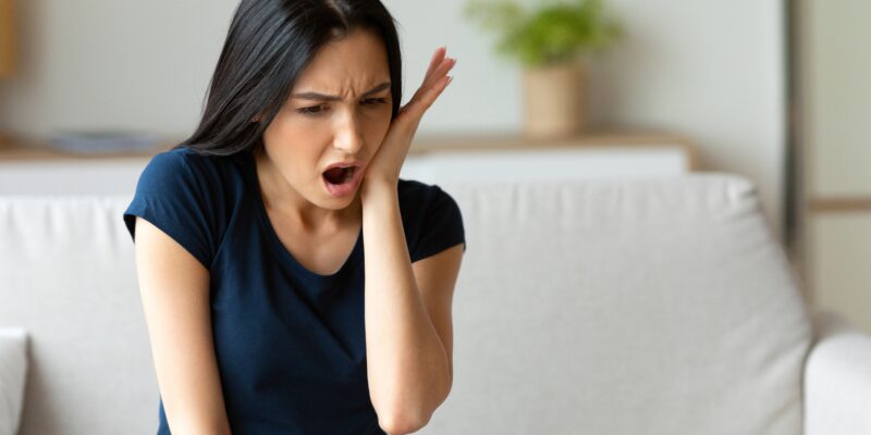 Toothache. Girl Suffering From Tooth Pain Touching Cheek Sitting On Couch At Home